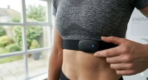 A woman in a UK home gym adjusting a black wireless heart rate chest strap over her workout top to sync with a cross trainer.